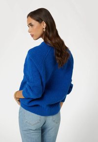 Niebieski sweter wykonany na drutach o prążkowanej fakturze, charakteryzujący się luźnym krojem i opadającymi ramionami, zestawiony z jasnoniebieskimi dżinsami.