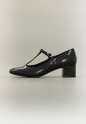 Chaussure Mary Jane en cuir verni noir avec un talon bloc bas et une bride en T avec boucle ronde sur un fond neutre.