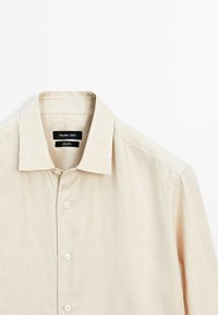 Camicia in lino beige con colletto a punta e chiusura frontale a bottoni. L'etichetta mostra "Massimo Dutti" con le istruzioni per la cura sotto. Texture morbida.