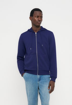 Mladi moški temne polti, oblečen v mornarsko modro prešito jopico z zadrgo in svetlo modre kavbojke, stoji z eno roko v žepu.