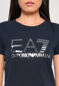Mørkeblå t-skjorte med korte ermer som har en metallisk sølv "EA7 Emporio Armani" logo, utsmykket med nagler og striper.