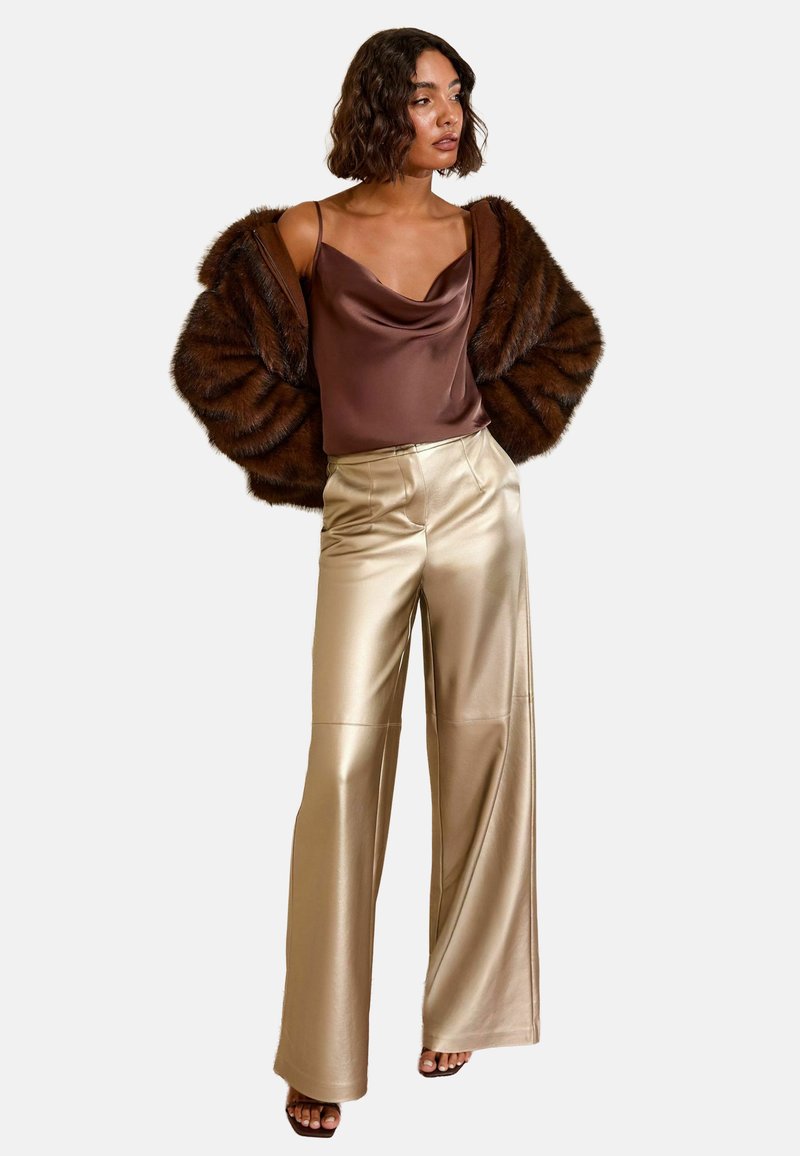 Hnědý umělý kožich, saténový hnědý top a lesklé zlaté kalhoty širokého střihu. Outfit má hladké textury a uvolněný střih.