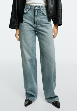 Jean taille haute, jambes larges bleu clair, porté avec des chaussures pointues noires, un top blanc et une veste en cuir noire.