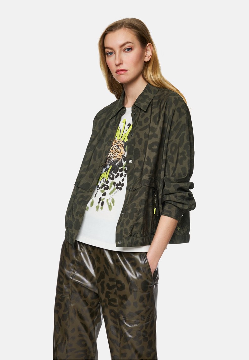 Leopard-patterned green jacket with a relaxed fit, paired with a white graphic tee featuring a leopard print, and shiny dark-colored trousers.