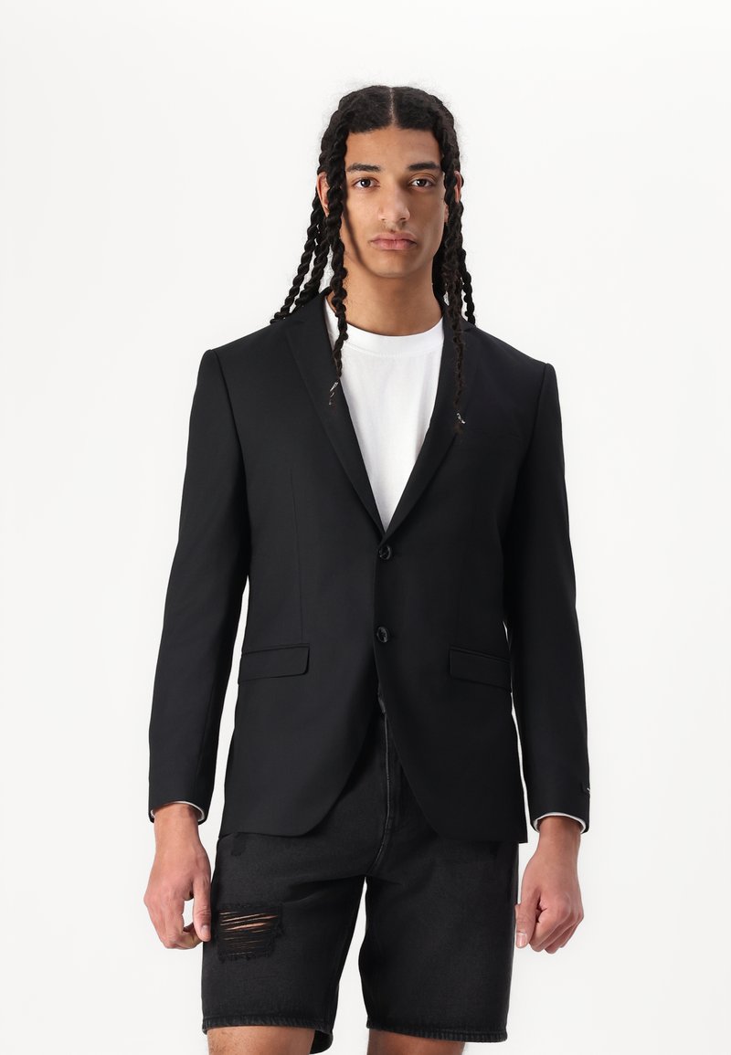 Sort blazer med to knapper, spidse revers og frontlommer, parret med en hvid t-shirt og sorte slidte shorts. Glat tekstur.