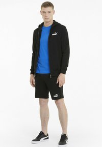 Felpa nera con zip sopra una t-shirt blu, abbinata a pantaloni corti neri e sneakers nere. Presenta loghi sugli indumenti. Design pulito.