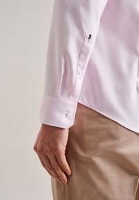 Helles rosa Hemd mit tailliertem Schnitt, Knopfmanschetten und subtiler schwarzer Rosenstickerei an den Ärmeln. Der Stoff wirkt glatt.
