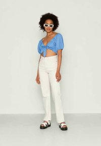 Blusa azul cropped com mangas bufantes, amarrada na frente; calças jeans brancas de cintura alta; sandálias chunky em preto e branco; óculos de sol brancos.