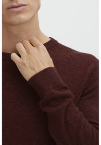 Burgundy knit sweater with a crew neckline, ribbed cuffs, and a soft texture, partially shown with a hand touching the collar.