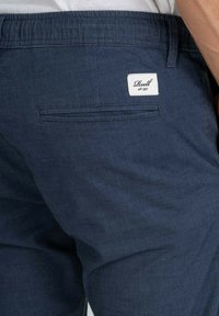 Navy blue shorts made from textured fabric, featuring a back pocket with a white label that has "Reell est. 1997" embroidered.