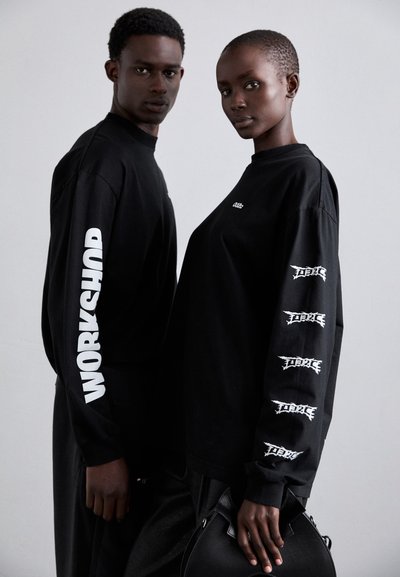 Black long-sleeve shirts feature white graphics on sleeves. One shirt reads "WORKSHOP," and the other has repeated logos. Black handbag visible.