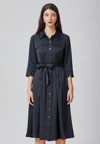 Robe noire à boutons, confectionnée dans un tissu lisse. Présente un corsage ajusté, une jupe fluide et une ceinture à nouer à la taille. Design de col classique.