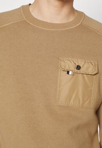 Beige gestrickter Pullover mit einem strukturierten Baumwollkörper und einer kontrastierenden Nylon-Tasche mit Knopfverschluss, quadratischer Form und geribtem Halsausschnitt.