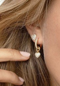 Orecchini in tono oro con perni a forma di cuore e un ciondolo a cuore pendente, decorati con strass trasparenti per un tocco di brillantezza.