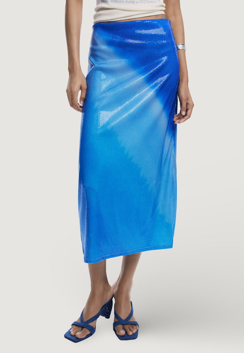 Midi-Rock aus blauen Pailletten mit Gradientendesign, glatter Textur und Wickelsilhouette. Kombiniert mit blauen Riemchenschuhen.