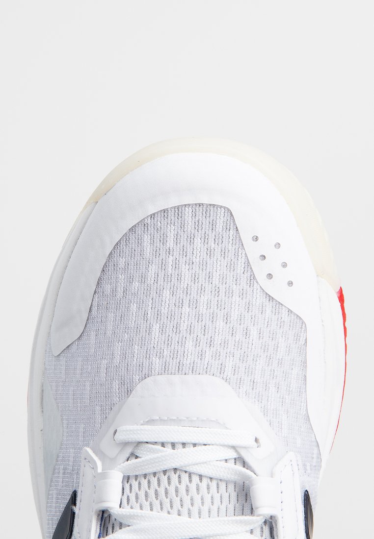 Zapatilla deportiva blanca con parte superior de malla gris texturizada, cuello acolchado y acento rojo en el talón. Presenta cordones blancos y orificios de ventilación en los lados.