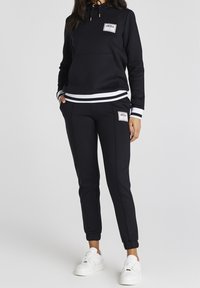 Černá mikina a odpovídající teplákové kalhoty vyrobené z měkké látky. Obsahuje bílé detaily, přední kapsu a značkové nášivky. Bílá obuv dokončuje vzhled.