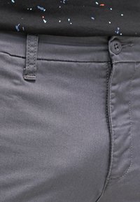 Grijze broek van gladde stof, met een knoopsluiting en standaard voorzakken. Subtiele stikseldetails zichtbaar.