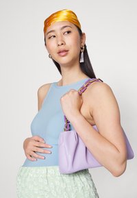 Bolsa lilás com uma forma estruturada, apresentando uma alça de corrente dourada e roxa; modelo usa uma t-shirt de alças azul com nervuras e uma saia texturizada em menta.