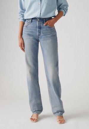 Personne portant un jean en denim bleu clair et une chemise bleu clair boutonnée, une main dans la poche et des sandales beiges à bout ouvert.