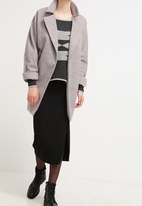 Personne portant un manteau gris ouvert sur le devant, un pull à motif géométrique noir et gris, une jupe midi noire fendue, des collants noirs et des bottes noires.