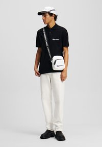 Μαύρο πόλο μπλουζάκι με κοντά μανίκια, λευκή επωνυμία, λευκή τσάντα που φοριέται διαγώνια, ανοιχτόχρωμο παντελόνι, μαύρα παπούτσια, λευκό καπέλο με λεπτομέρειες.