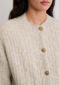 Cremefarbener Cardigan mit Zopfmuster aus weichem Material, mit rundem Ausschnitt und drei großen, hellbraunen Knöpfen auf der Vorderseite.