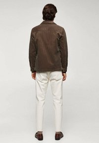 Uomo che indossa una giacca in corduroy marrone, pantaloni bianchi e scarpe marroni, in piedi e rivolto di spalle su uno sfondo chiaro e semplice.