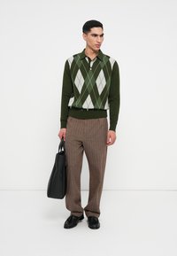 Grünes Polo mit Argyle-Muster, braune gestreifte Hose und schwarze Lederschuhe. Modell hält eine schwarze Tragetasche. Neutraler Hintergrund.
