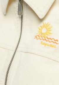 Veste couleur crème avec fermeture éclair argentée, ornée d'un soleil jaune et de vagues orange brodées près du col avec le texte "Icewear".