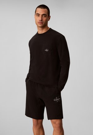 Hombre con camisa negra de manga larga y pantalones cortos con logos de Calvin Klein, de pie con las manos en los bolsillos contra un fondo gris liso.