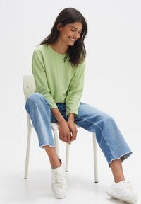 Hellgrünes geripptes Langarmoberteil, kombiniert mit hellblauen Cuffed-Jeans. Das Modell trägt weiße Sneaker und sitzt auf einem Stuhl vor einem neutralen Hintergrund.