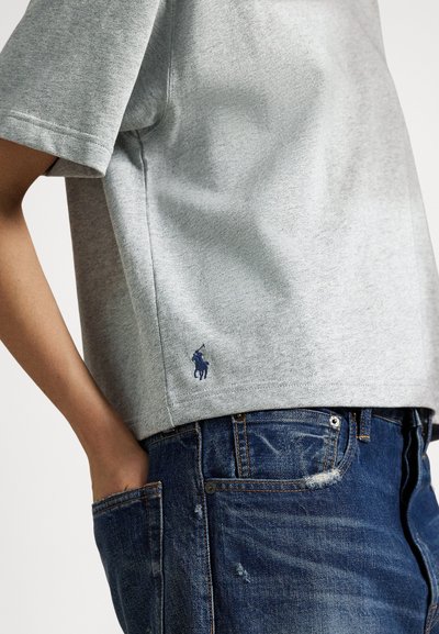 Osoba nosi svetlosivú krátku košeľu s malým logom hráča póla a modré opotrebované džínsy s rukou v vrecku.