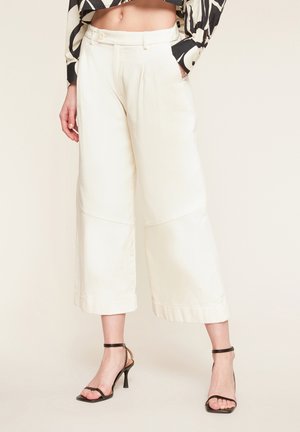 Woman wearing high-waist wide-leg white cropped pants paired with black strappy high-heeled sandals, one hand in pocket.