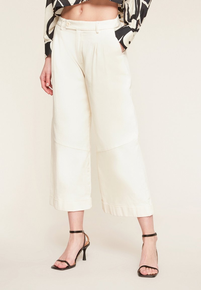 Femme portant un pantalon blanc à jambes larges et taille haute, raccourci, associé à des sandales noires à talons hauts et fines lanières, une main dans la poche.