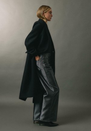 Schwarzer langer Mantel mit Puffärmeln, kombiniert mit glänzenden grauen weit geschnittenen Jeans und spitzen schwarzen Absätzen; Seitenprofil vor einem grauen Hintergrund.