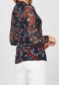 Genomskinlig marinblå blus med röd, blå och vit blommönster, slim fit, långa ärmar och knäppta ärmslut, kombinerad med vita byxor.