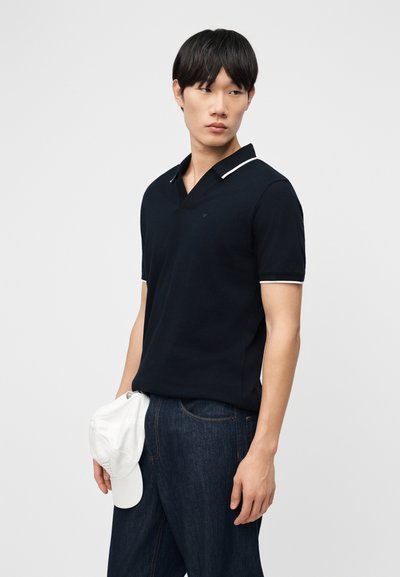 Άνδρας με μπλε πόλο πουκάμισο και σκούρο τζιν, κρατώντας ένα λευκό καπέλο μπέιζμπολ στο αριστερό του χέρι, κοιτάζει πλάγια μπροστά σε απλό φόντο.