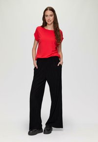 Rotes Kurzarmhemd kombiniert mit schwarzen, gefalteten, weiten Hosen. Die Hose hat seitliche Taschen. Die Schuhe sind schwarz mit einer flachen Sohle.