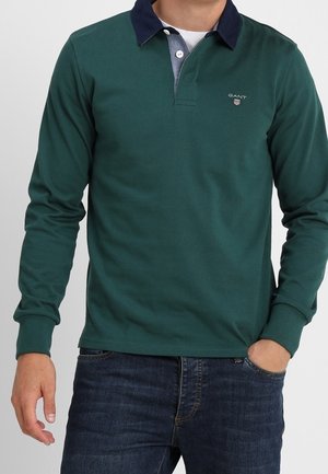 Hombre que lleva una camisa tipo polo verde oscuro de manga larga con cuello azul marino y vaqueros azules, de pie con una mano en el bolsillo.