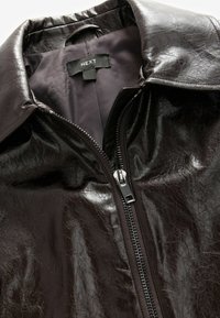 Veste en similicuir marron à col montant, fermeture éclair diagonale et texture lisse. L'intérieur présente une doublure foncée et une étiquette.