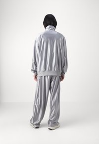 Osoba stojící zády ke kameře, oblečená v volném, leskl ém stříbrném teplákovém kompletu s vysokým límcem a bílými teniskami na jednobarevném bílém pozadí.