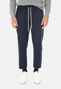 Uomo che indossa pantaloni jogger blu navy con cordini bianchi, calze nere, scarpe da ginnastica bianche e una maglietta grigia a maniche lunghe, in piedi contro uno sfondo bianco.