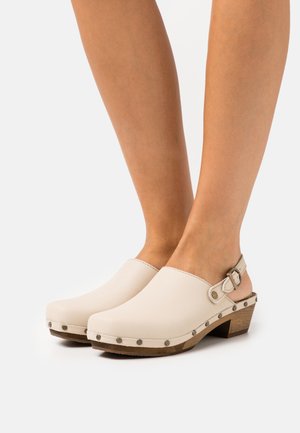 Crème leren clogs met een ronde neus, slingbackriem en decoratieve metalen studs langs de houten zool. Gladde textuur met een minimaal ontwerp.