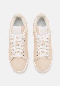 Sapatilhas de camurça bege com atacadores brancos, apresentando um colarinho almofadado e uma etiqueta branca com o logótipo na língua. Sola de borracha plana e costura mínima.