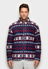 Giacca in pile blu navy e bianca con motivi geometrici rossi e bianchi; presenta un colletto alto e una cerniera frontale completa. Texture morbida, maniche lunghe.