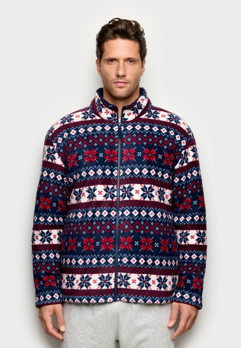 Giacca in pile blu navy e bianca con motivi geometrici rossi e bianchi; presenta un colletto alto e una cerniera frontale completa. Texture morbida, maniche lunghe.