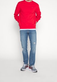 Sweatshirt rouge à col rond, associé à un jean léger bleu déchiré et des baskets grises avec des accents rouges. Tenue simple et décontractée.