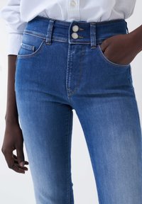 Jeans de mezclilla azul con una cintura alta, que cuentan con dos botones, bolsillos frontales y un desvanecimiento sutil cerca de los muslos. Textura suave y ajuste entallado.