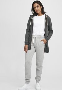 Grå zip-up hettegenser med snøring, lagd over en hvit T-skjorte. Lysegrå joggebukser med mansjetter, kombinert med hvite joggesko.